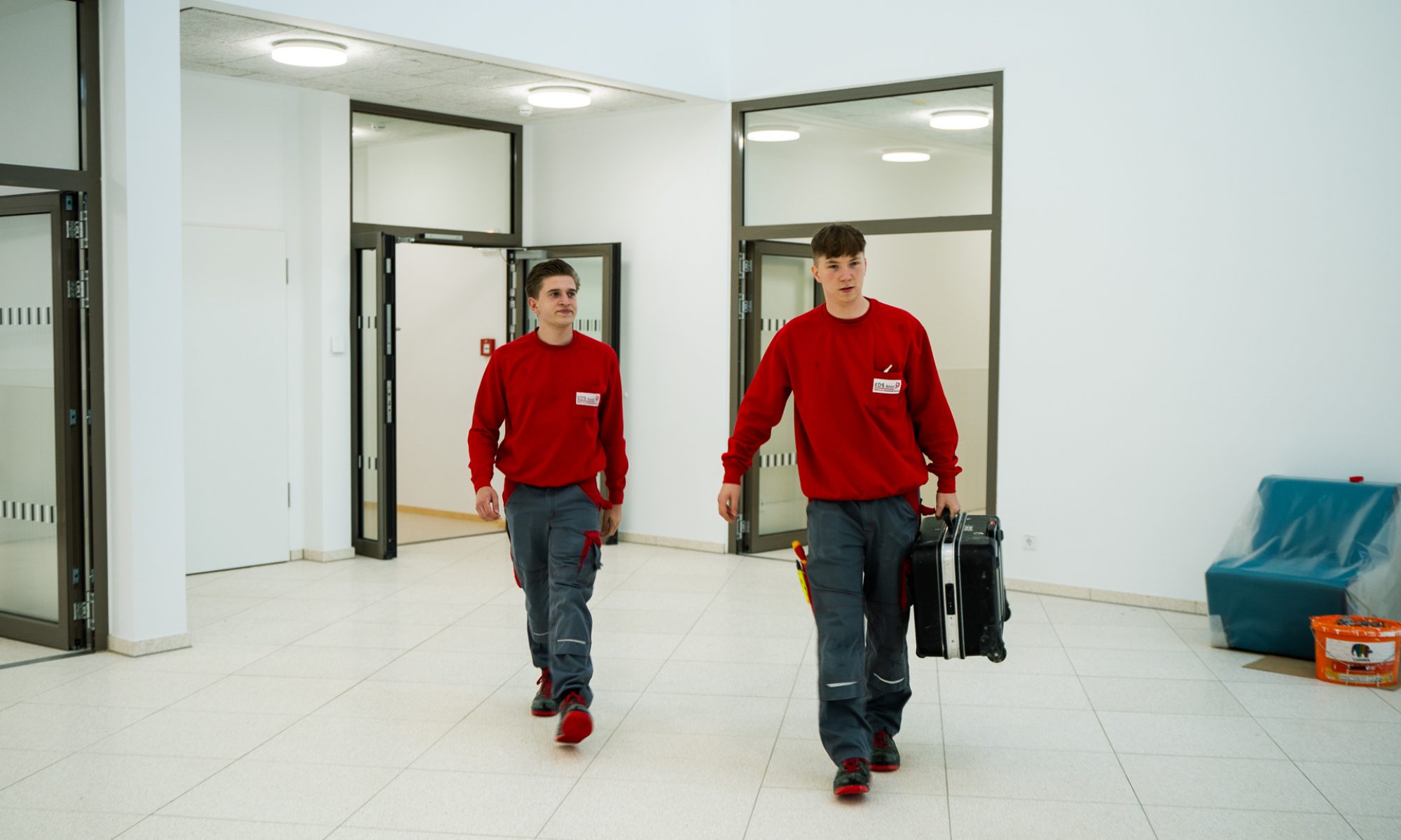 Zwei Mitarbeiter in roten Pullovern betreten mit einer Werkzeugtasche das Firmengebäude.